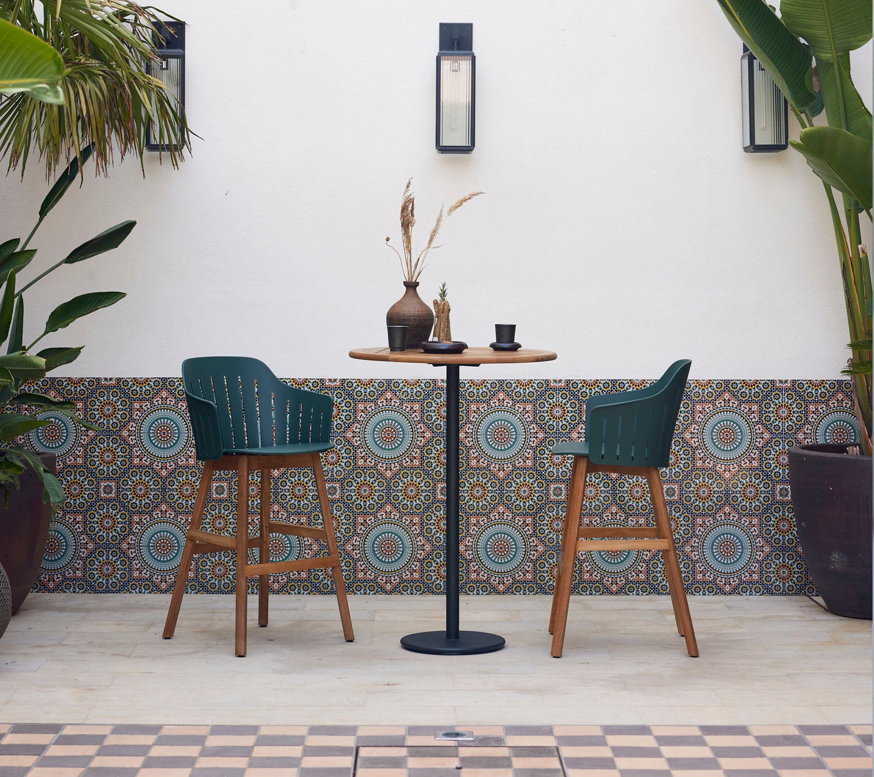 Een stijlvolle bartafel met twee groene stoelen, versierd met een vaas en een kop, omringd door geglazuurde tegels en weelderige planten.