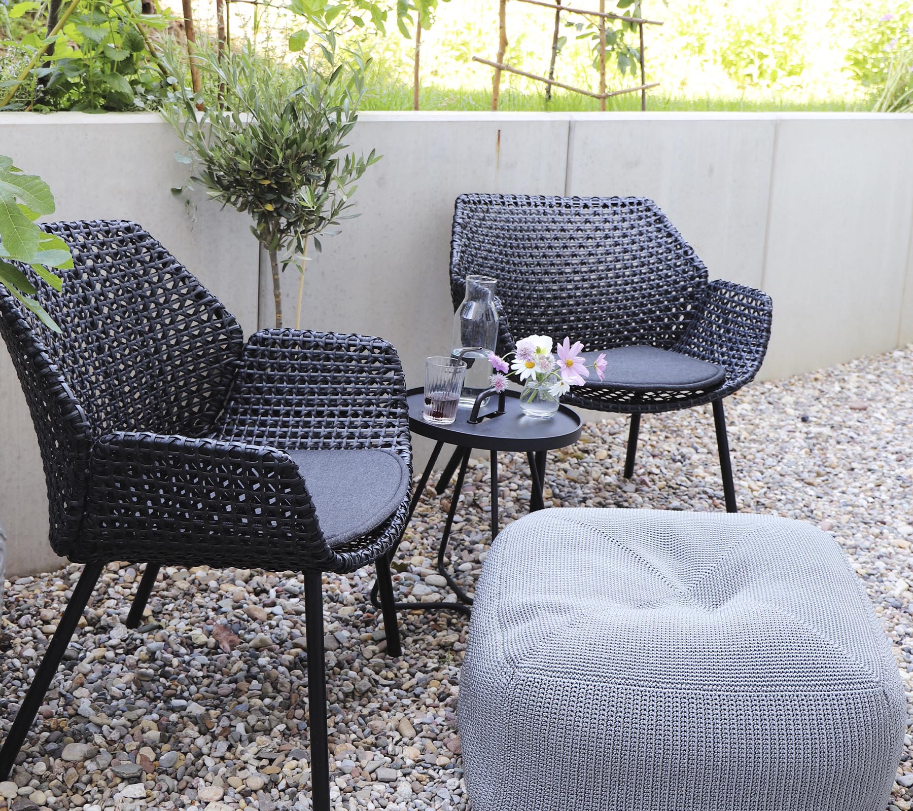 Twee stijlvolle zwarte stoelen in combinatie met een klein tafeltje, versierd met een vaas met bloemen en een gezellige poef op een grindoppervlak.