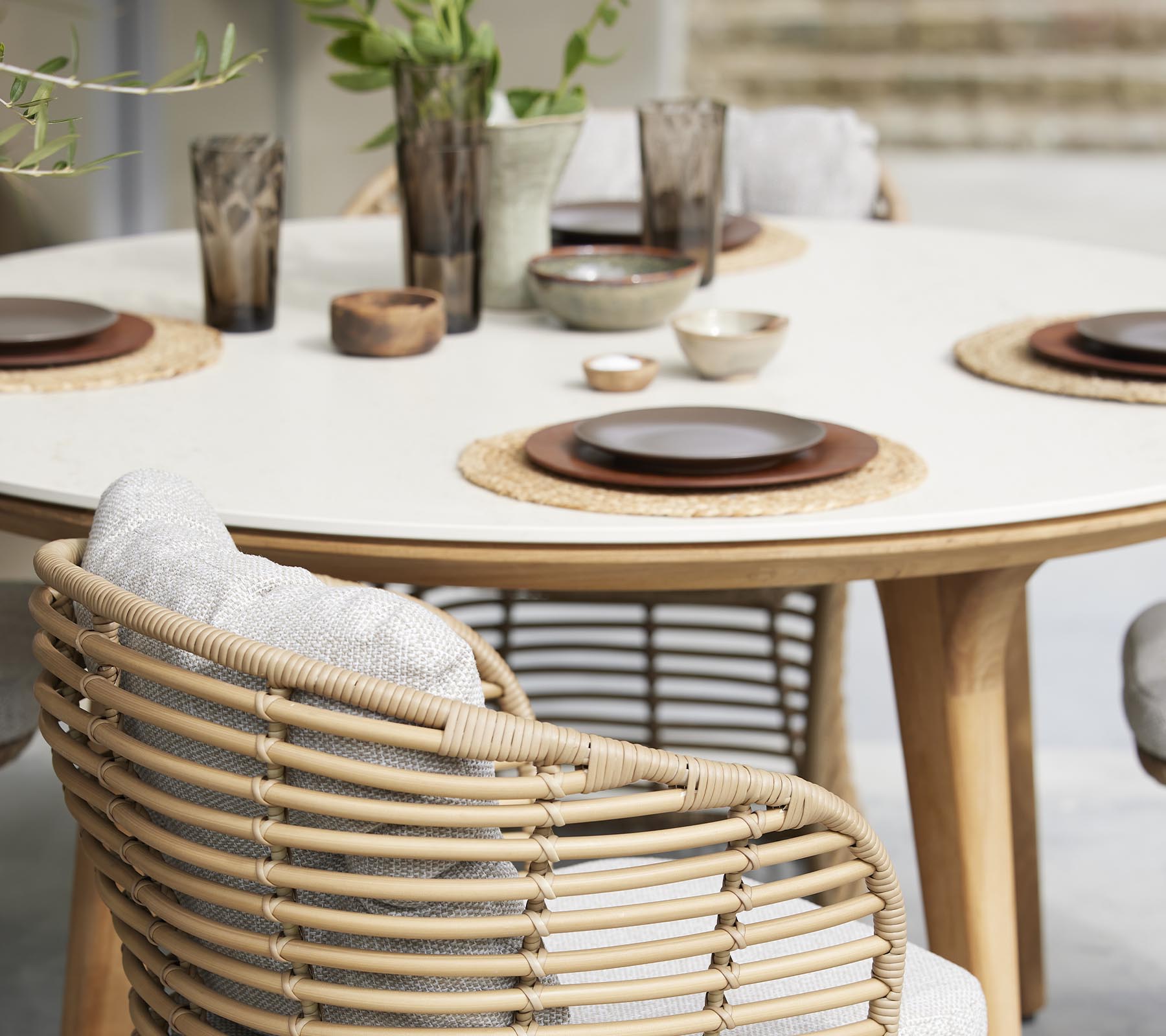 Een stijlvolle eettafelset met borden, kommen en decoratieve glazen, omringd door gevlochten stoelen en natuurlijke elementen.