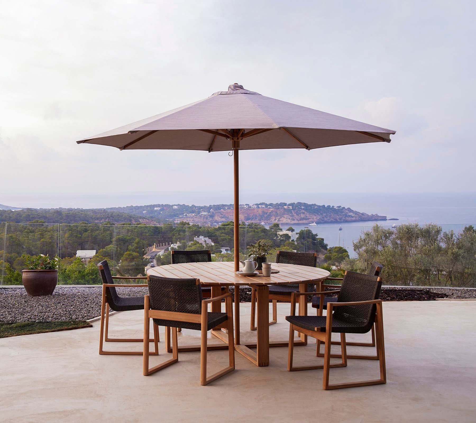 Een ronde houten tafel met zes stoelen onder een groot parasol, tegen een schilderachtige achtergrond van heuvels en de zee.