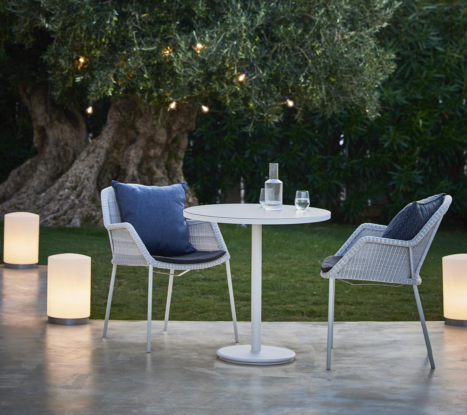 Een gezellige zithoek met twee moderne stoelen, een ronde tafel en stijlvolle lampen, omringd door groen en warme verlichting.