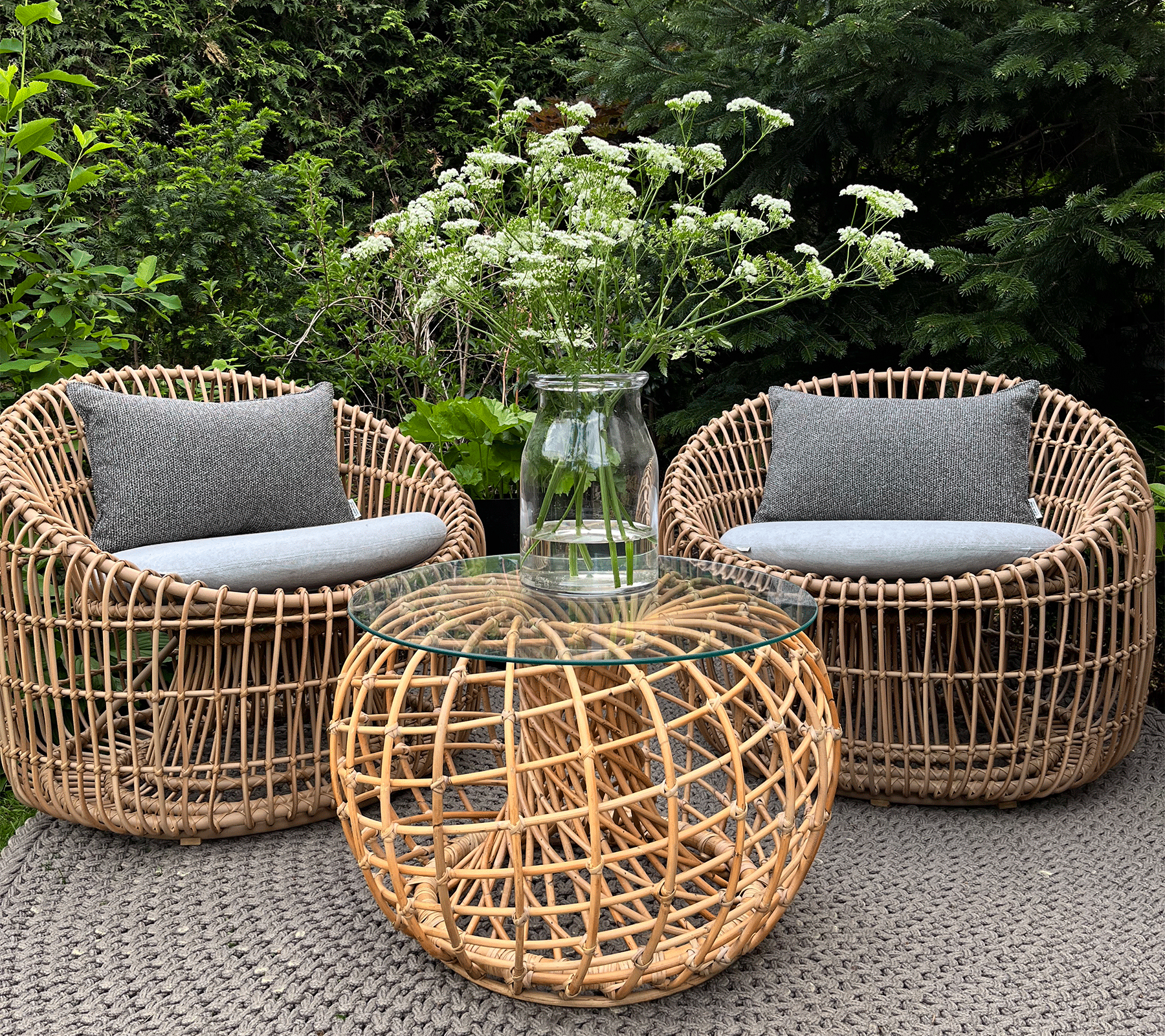 Twee geweven stoelen met grijze kussens omringen een rond geweven salontafel, bedekt met een heldere vaas met witte bloemen.