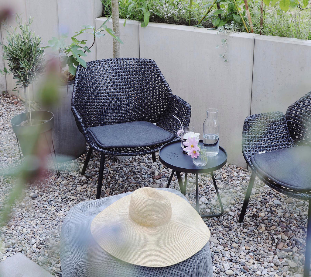 een gezellige zithoek met gevlochten stoelen, een klein tafeltje, een glas water, bloemen en een strohoed, omringd door groen en stenen.