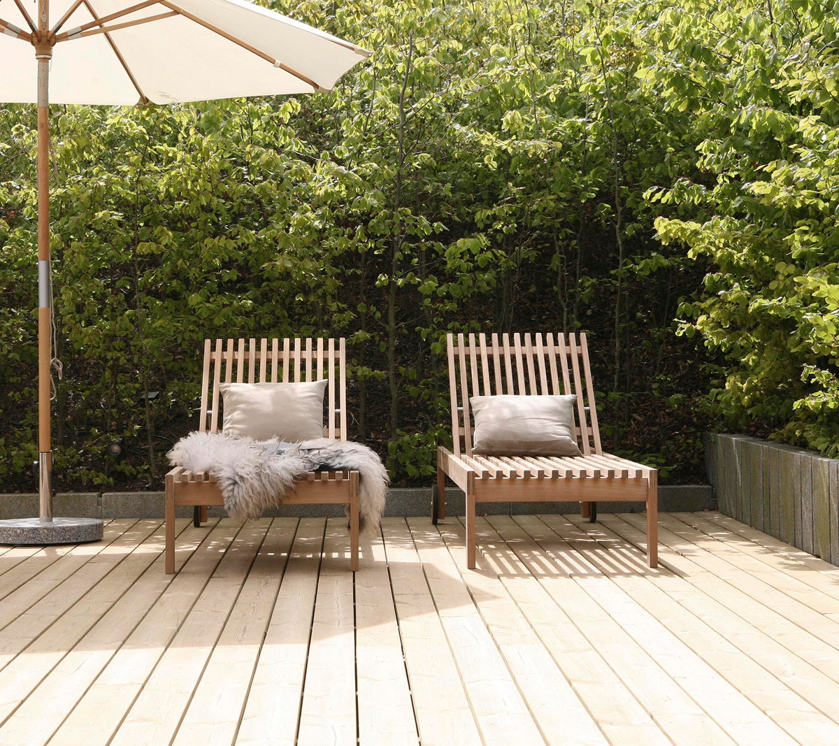 Twee stijlvolle houten lounge stoelen met kussens, geplaatst op een houten terras, omringd door groen en een grote parasol in de buurt.