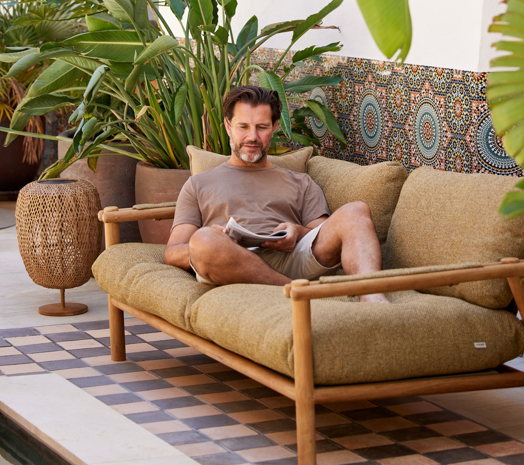 Een man ontspant op een comfortabele bank, leest een tijdschrift, omgeven door weelderig groen en decoratieve patronen.