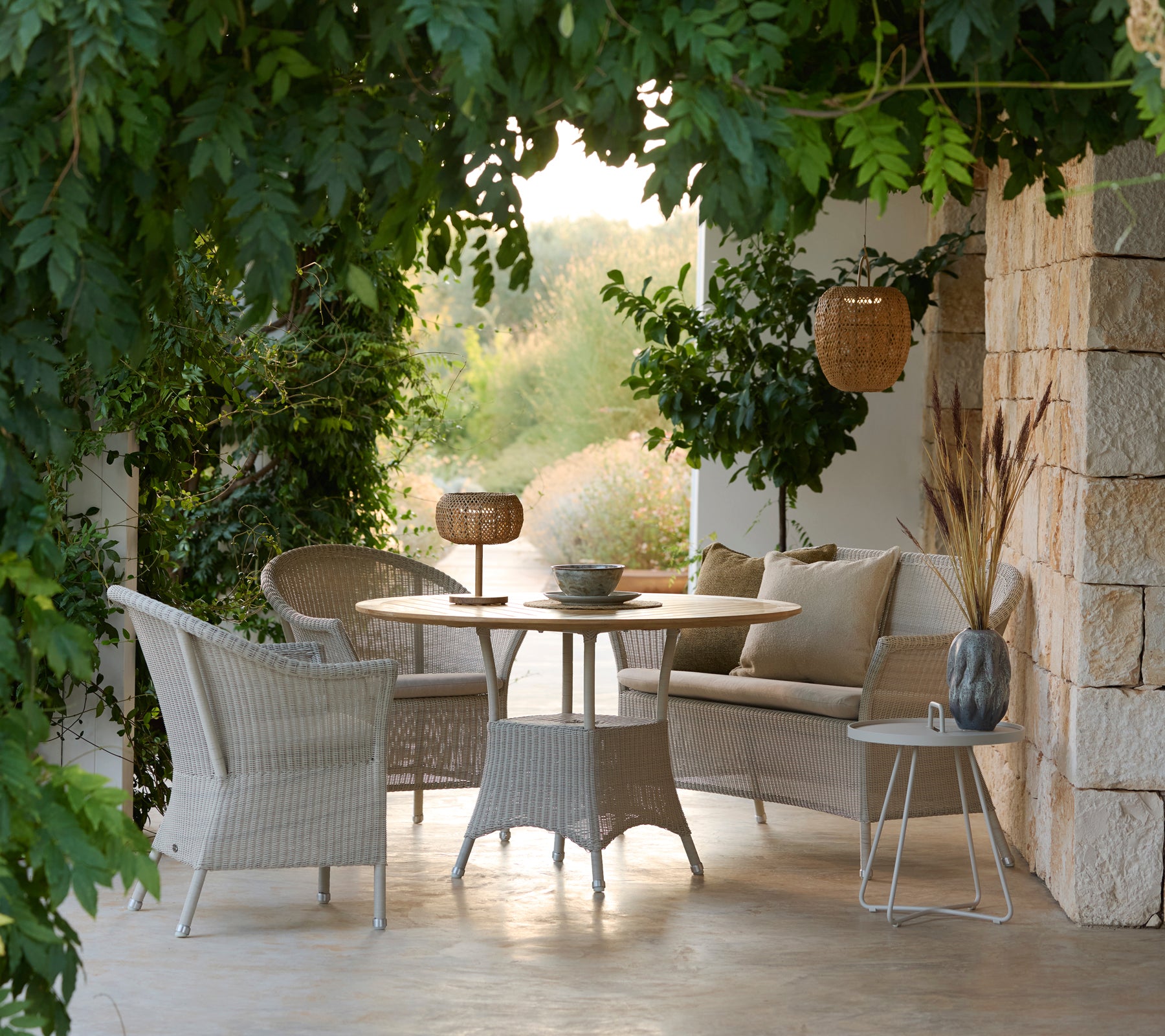 Tijdloze loungebank set met gevlochten stoelen, ronde tafel en kussens in een serene buitenomgeving.