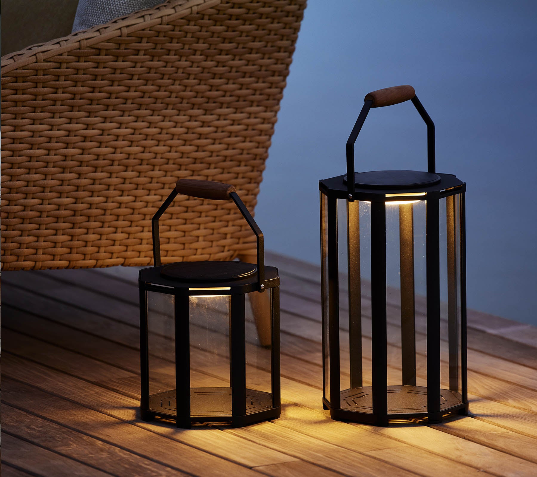 Twee stijlvolle Lightluxe-laternen met glazen panelen en handvatten, die een warme gloed uitstralen, naast een geweven stoel geplaatst.