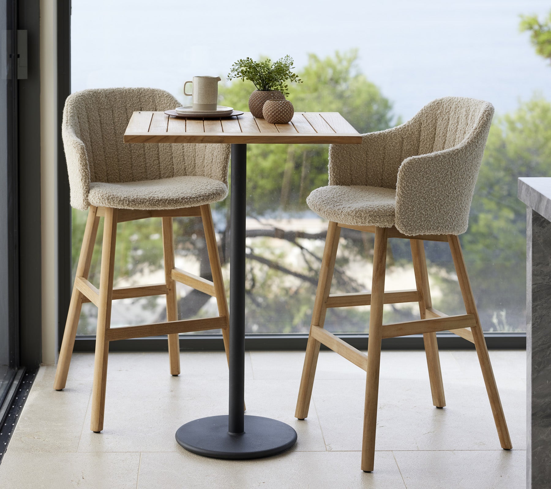 Een kleine tafel met twee gestoffeerde stoelen, een kop en decoratieve items, tegen een natuurlijke achtergrond.