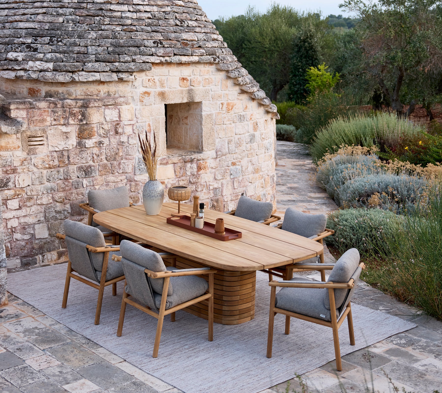Houten eettafel met grijze kussensstoelen in een rustieke buitenomgeving.