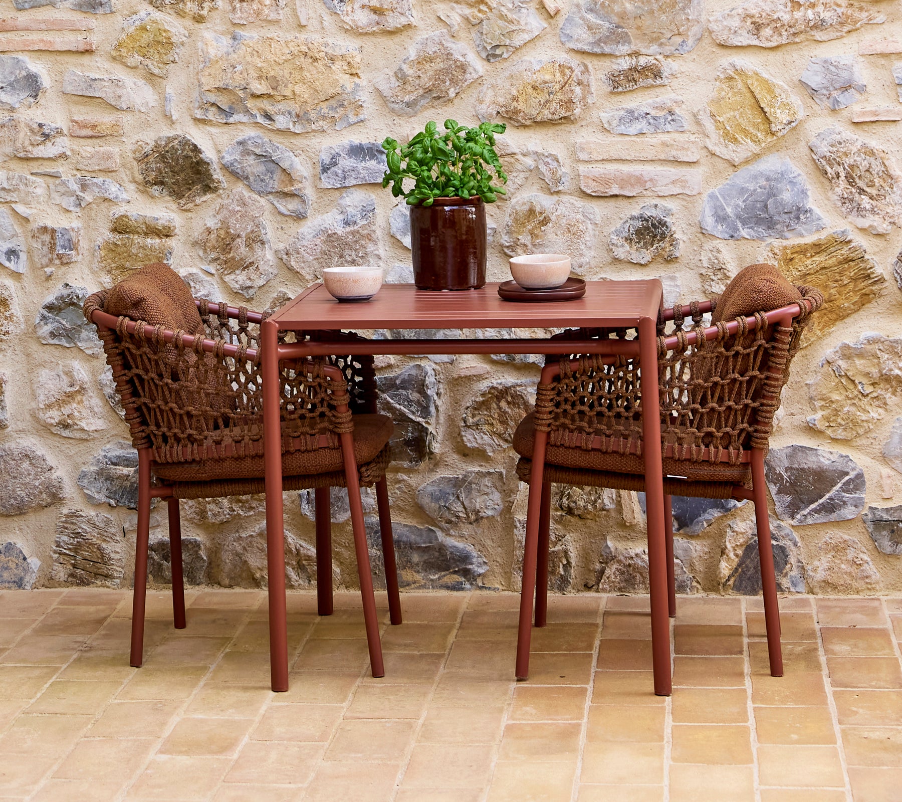 Bruine ombre stoelen gecombineerd met een bijpassende tafel, geaccentueerd door een kamerplant en decoratieve kommen, tegen een stenen muur.