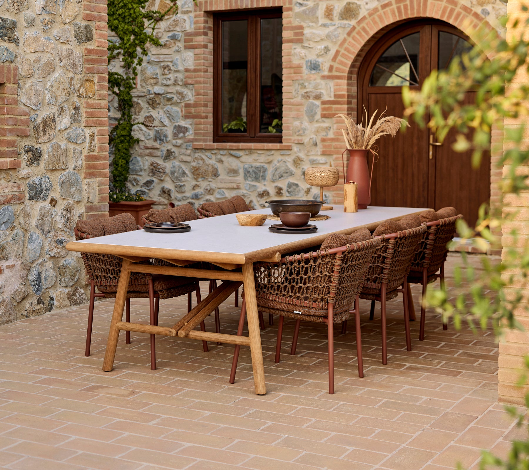 Buiten eetopstelling met umber bruine Ocean eetstoelen rond een teak Sticks eettafel, tegen een stenen muur.