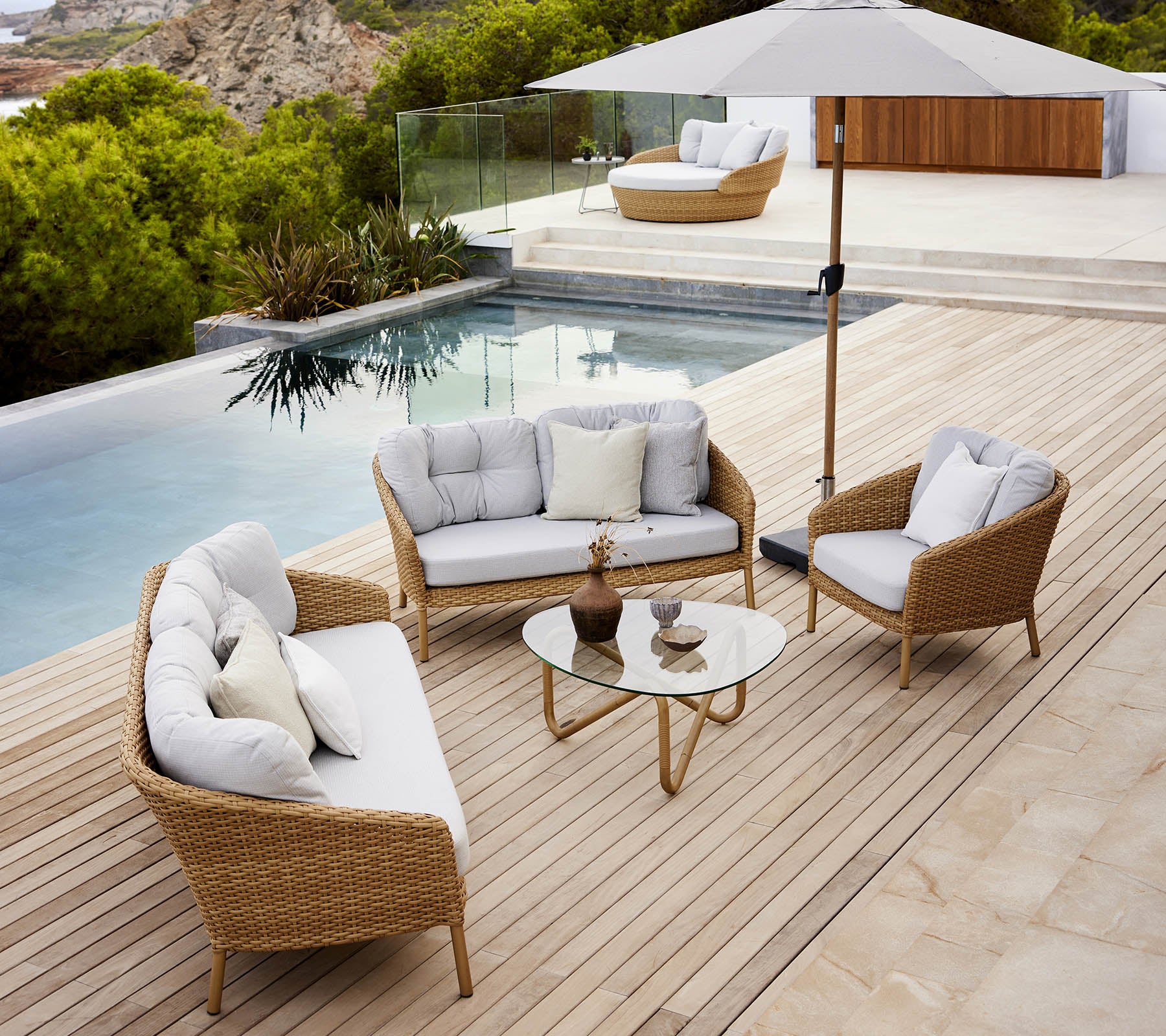 Moderne buitenlounge met een glazen tafel, comfortabele zitplaatsen en een parasol aan de rand van het zwembad met weelderig groen.