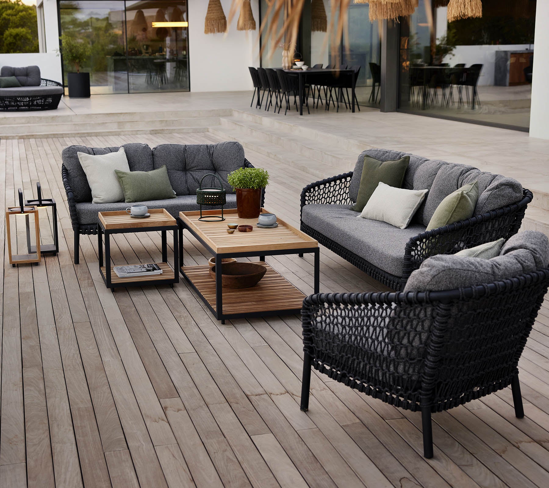 Stijlvol buitenloungegebied met zwarte rotan meubels, grijze kussens, een houten salontafel en potplanten op een houten terras.