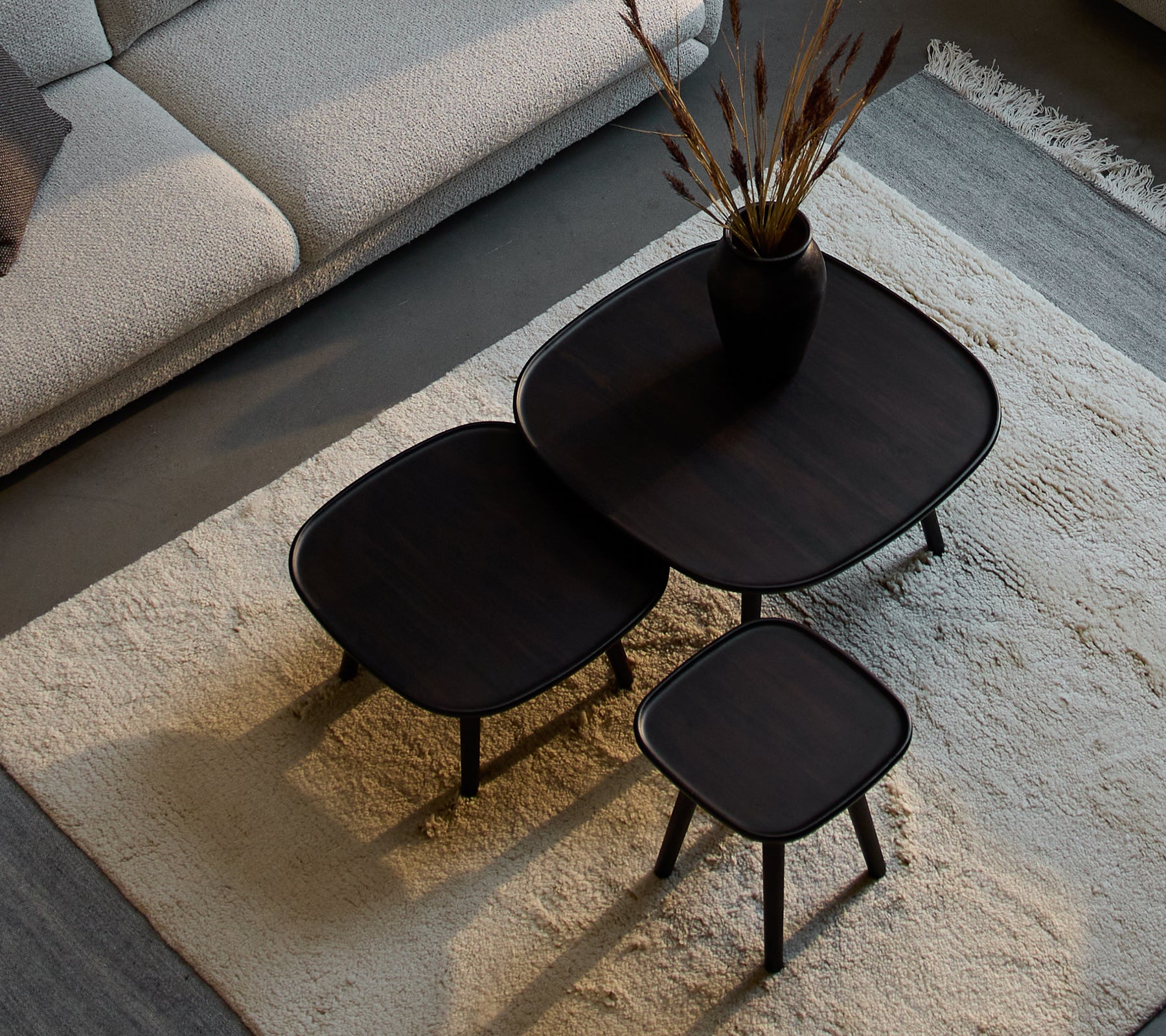 Drie stijlvolle Pace salontafels in verschillende maten, met een sleek ontwerp en donkere afwerking, vergezeld van een decoratieve vaas en gedroogde planten.