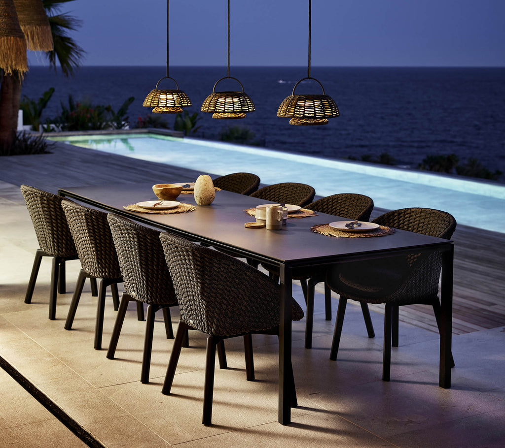Een stijlvolle dinersetting heeft een lange tafel omringd door pauwstoelen, verlicht door hanglampen boven een sereen uitzicht op het water.