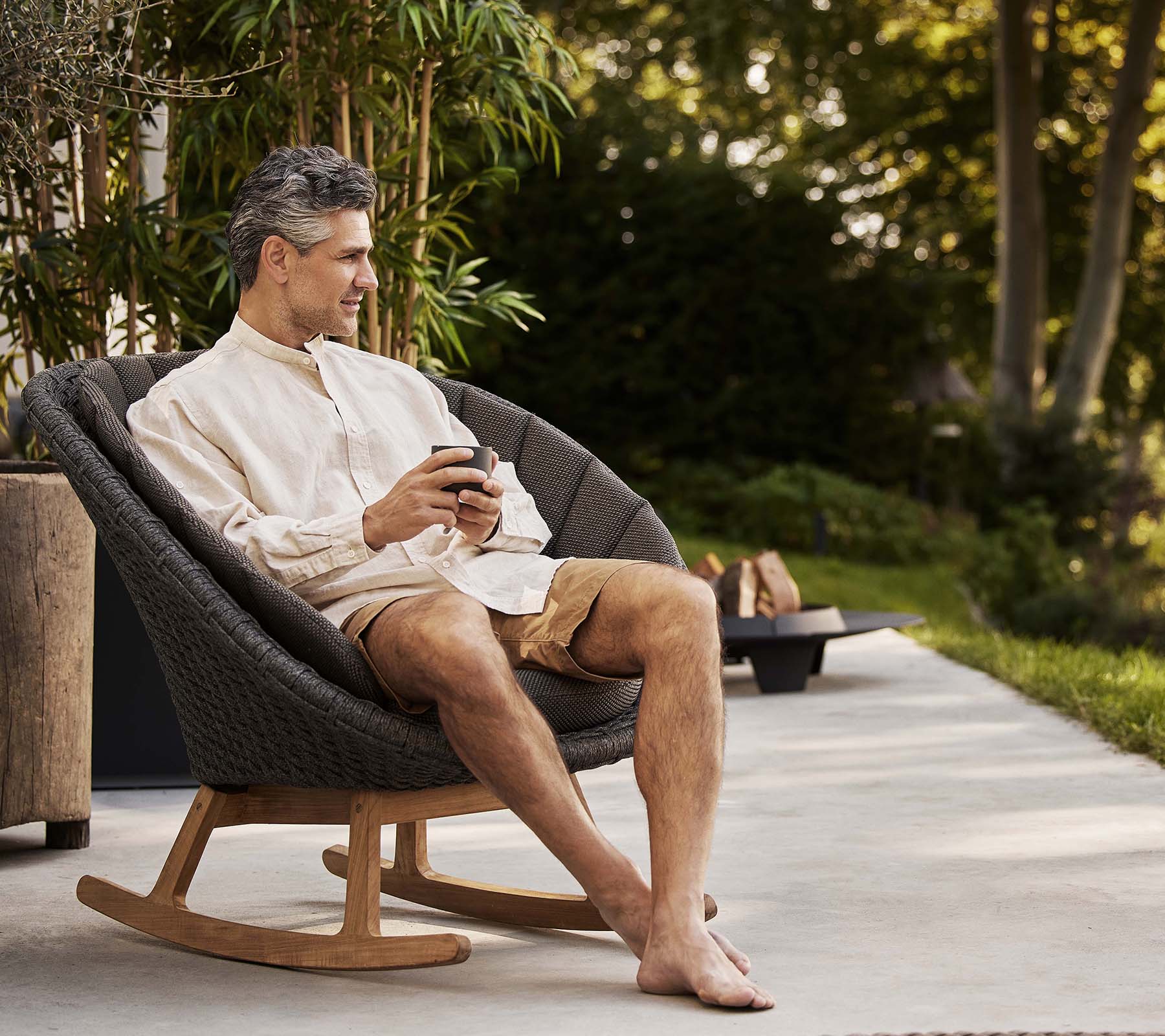Een man zit comfortabel in een stijlvolle pauwen schommelstoel en geniet van een moment van ontspanning terwijl hij een telefoon vasthoudt. Groene planten zijn op de achtergrond.