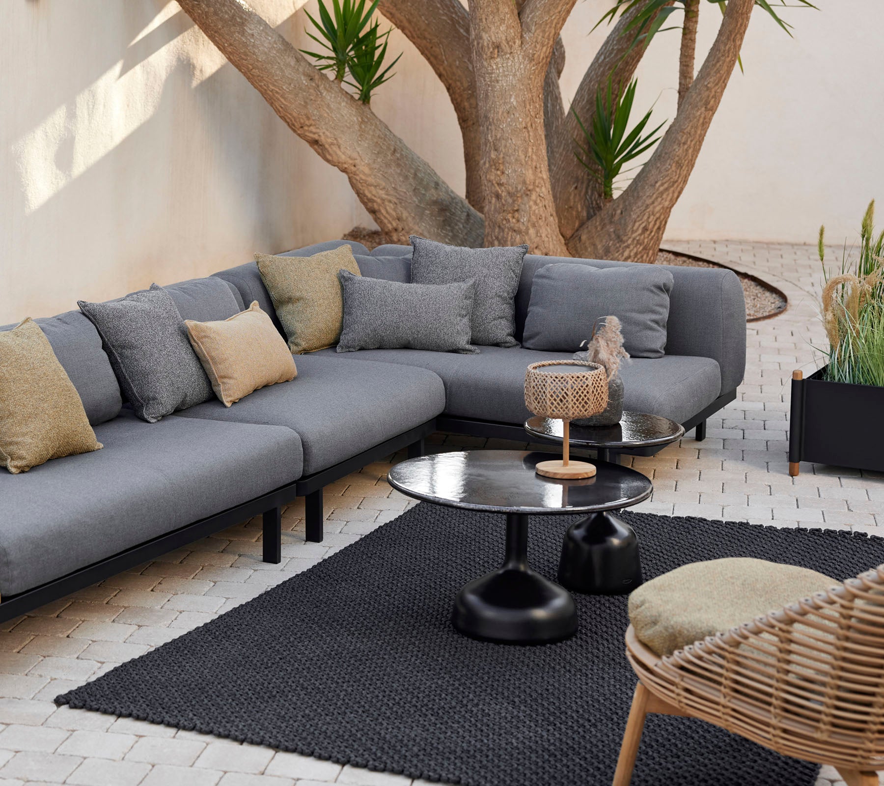 Loungegebied met een grijze modulaire sofa met decoratieve kussens, een strak salontafel en een textuurkleed omringd door groen.