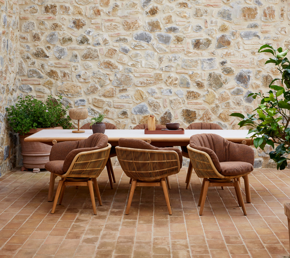 Een houten eettafel omringd door stijlvolle stoelen met teak poten, geplaatst tegen een gestructureerde stenen muur en versierd met groene planten.