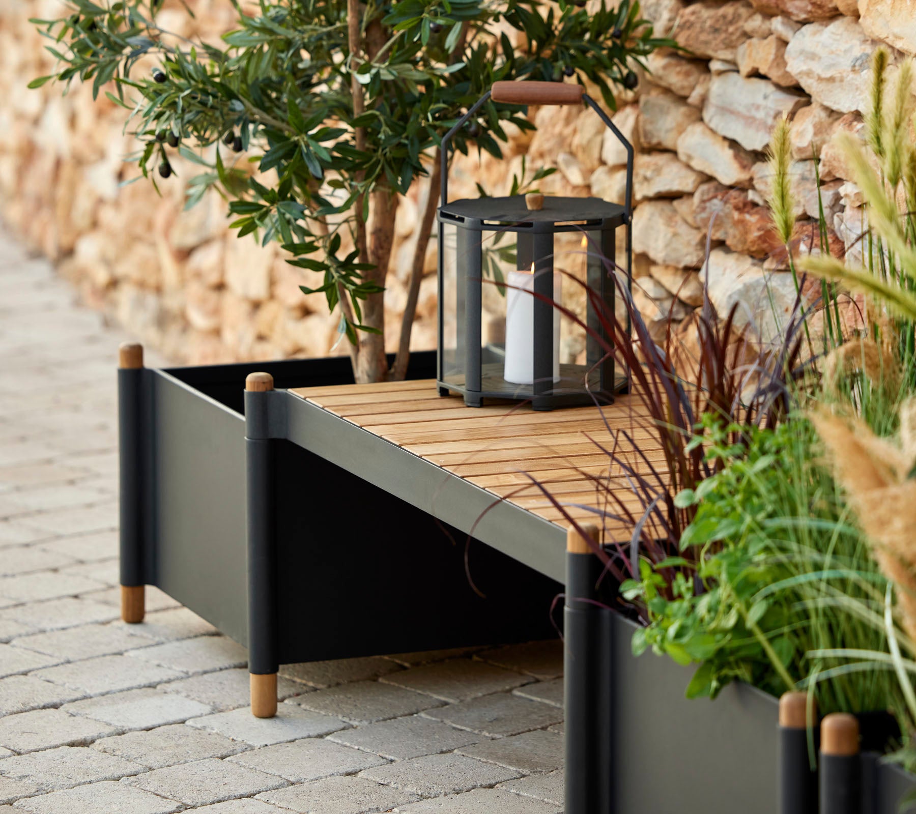 Een stijlvolle plantenbak met houten latten, omgeven door groen en een lantaarn, die een vleugje elegantie aan de ruimte toevoegt.