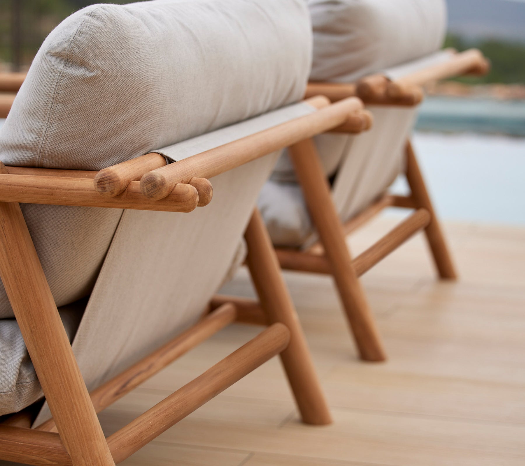 Twee stijlvolle loungestoelen met teak frames en comfortabele beige kussens, die een uniek stokontwerp op de rugleuning laten zien.
