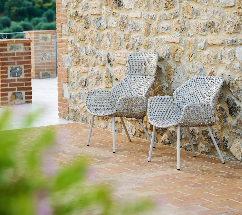 Twee Vibe hoge loungechairs in donker zand, stijlvol ontworpen, voor een gestructureerde stenen muur met een warme, uitnodigende sfeer.