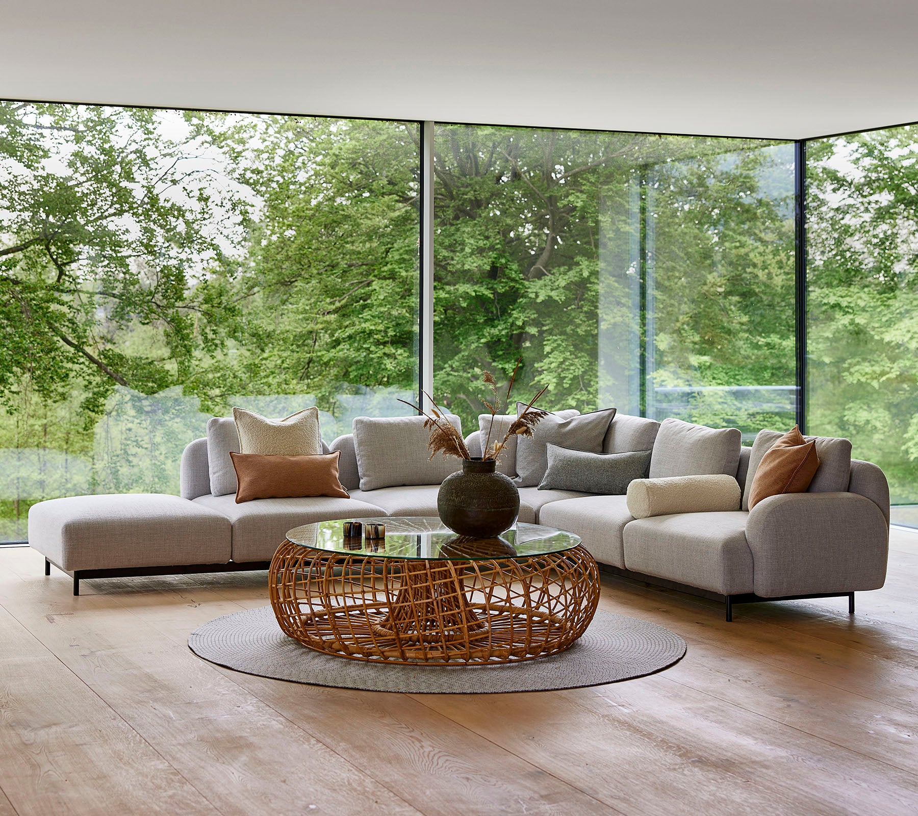 Moderne loungegebied met een modulair bankstel met decoratieve kussens, een ronde gevlochten salontafel en grote glazen ramen die groenvoorzieningen tonen.