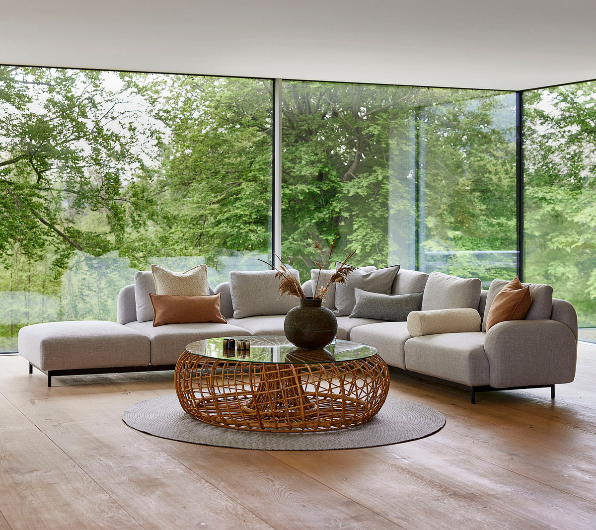 Moderne loungegebied met een modulair bankstel met decoratieve kussens, een ronde gevlochten salontafel en grote glazen ramen die groenvoorzieningen tonen.