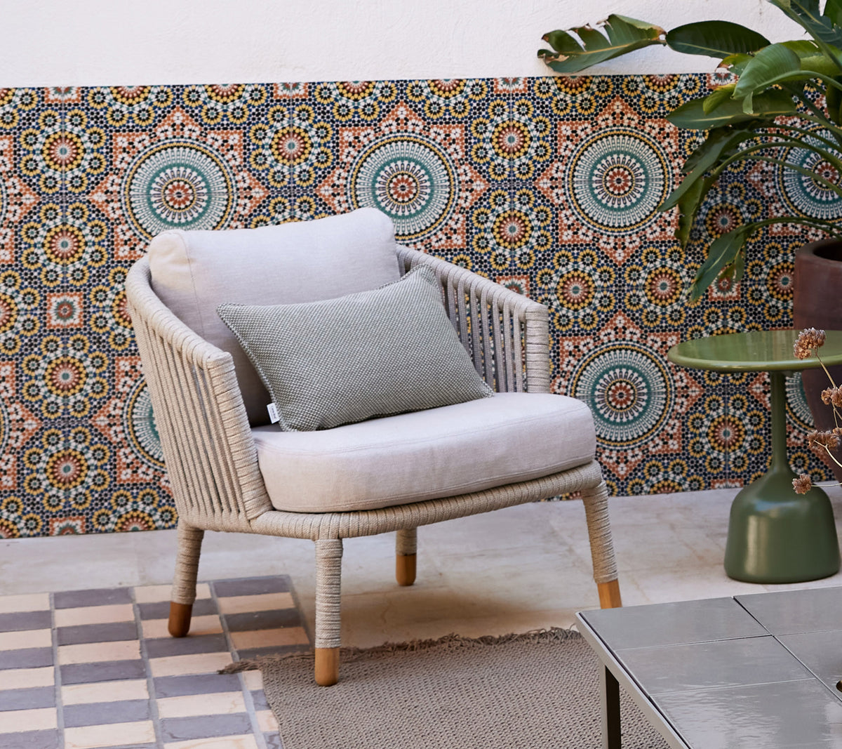 Een stijlvolle loungechaise met een zachte kussen en een decoratiekussen, geplaatst tegen een gemusterde achtergrond en omringd door een potplant.