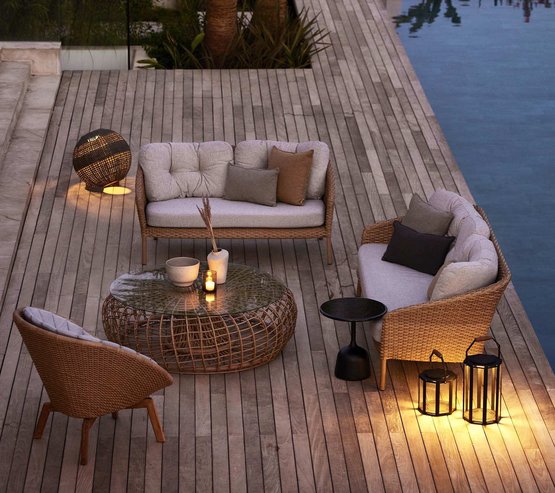 Gezellig buitensitgebied met gevlochten meubels, zachte kussens en sfeerverlichting naast een zwembad op een houten terras.