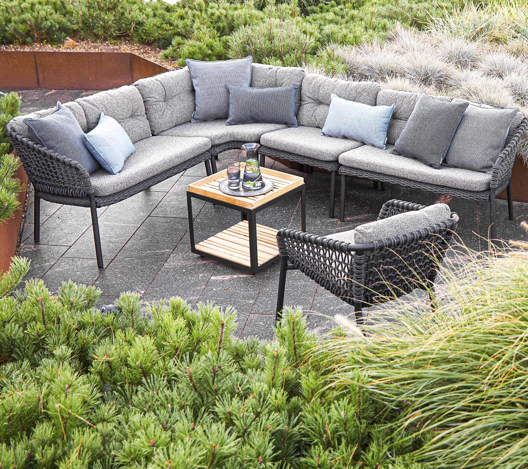 Stijlvolle buitenzitarrangement met een sectie sofa met kussens en een centraal tafel, omringd door groen.